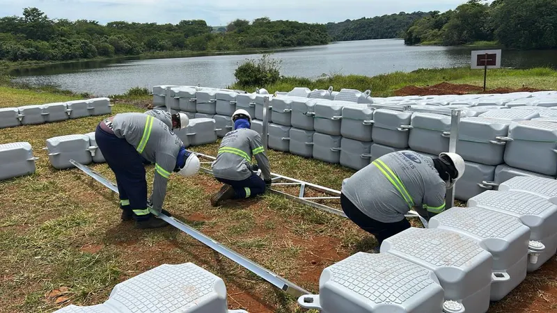 Três instaladores de colete montam a estrutura sobre os flutuadores empilhados na margem do reservatório