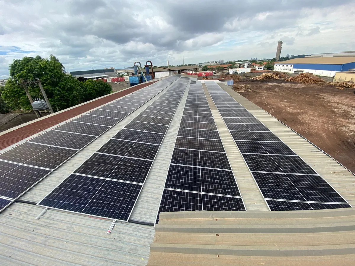 Fileiras de painéis solares no telhado metálico da Serraria Cidade Azul, com o pátio de toras ao fundo