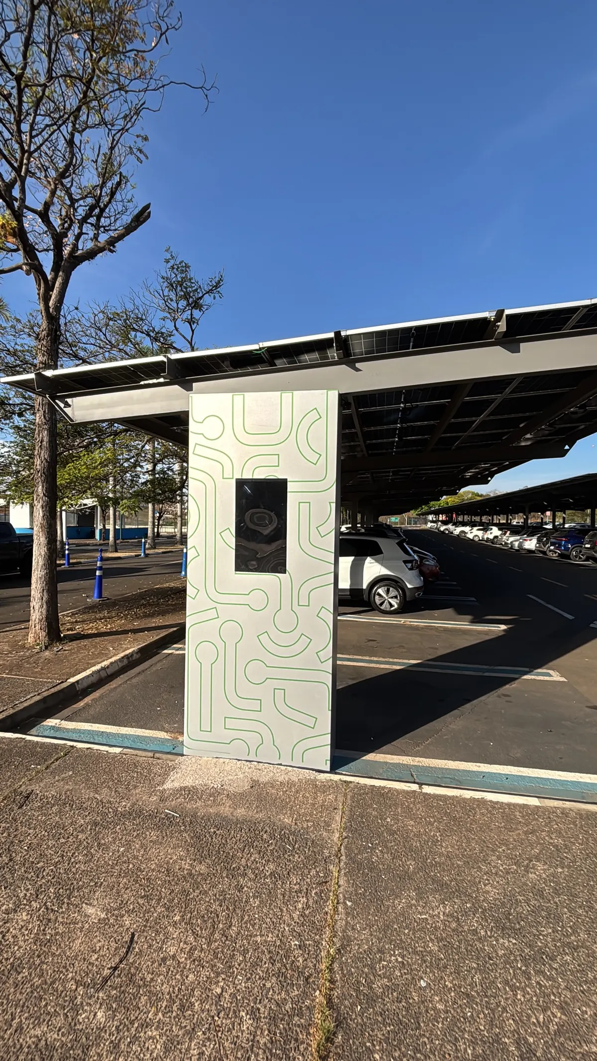Carregador de carro elétrico fixado no pilar do carport, com a cobertura de painéis solares acima e o estacionamento ao fundo