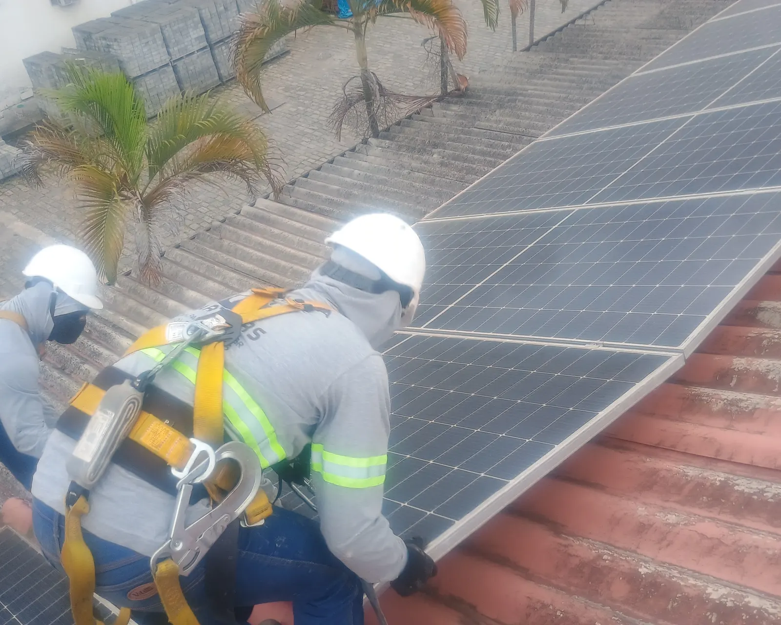 Dois técnicos de capacete, cinto paraquedista e talabarte trabalhando sobre um telhado de telha cerâmica com módulos solares