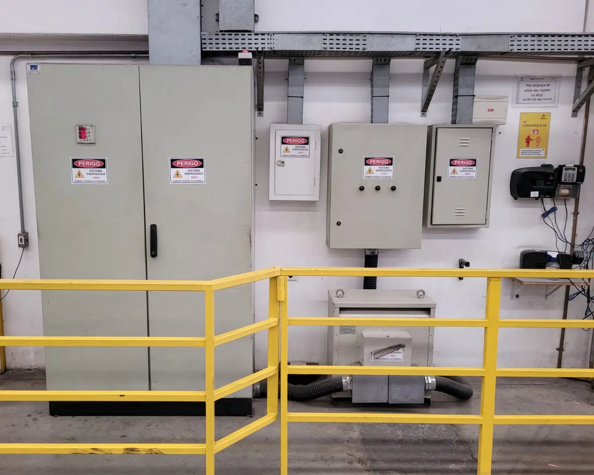 Quadros elétricos de uma fábrica com placas de perigo de sistema energizado, eletrocalha acima e guarda-corpo amarelo à frente